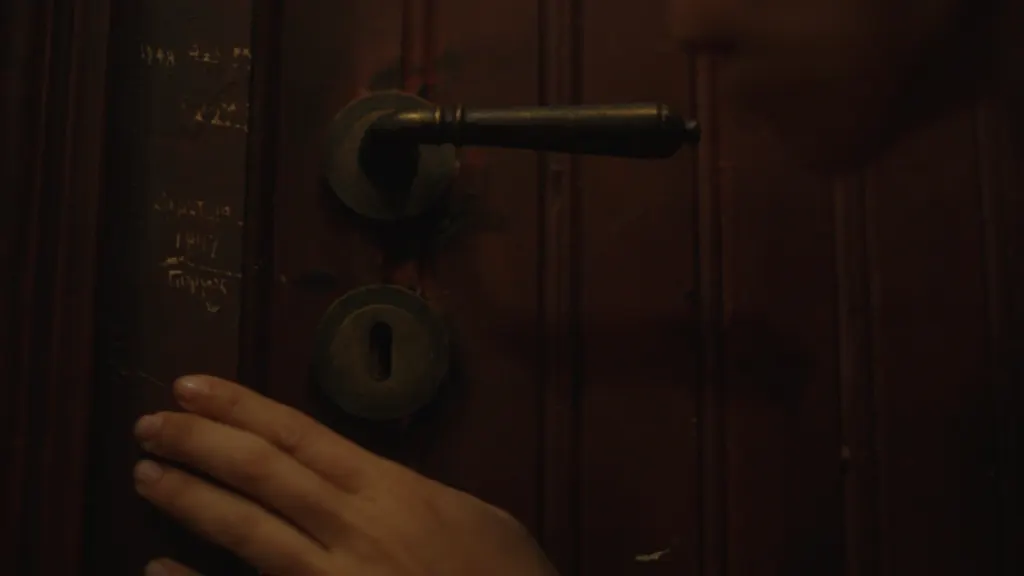 A hand on an old wooden door with a brass handle and Greek writing scratched into the frame